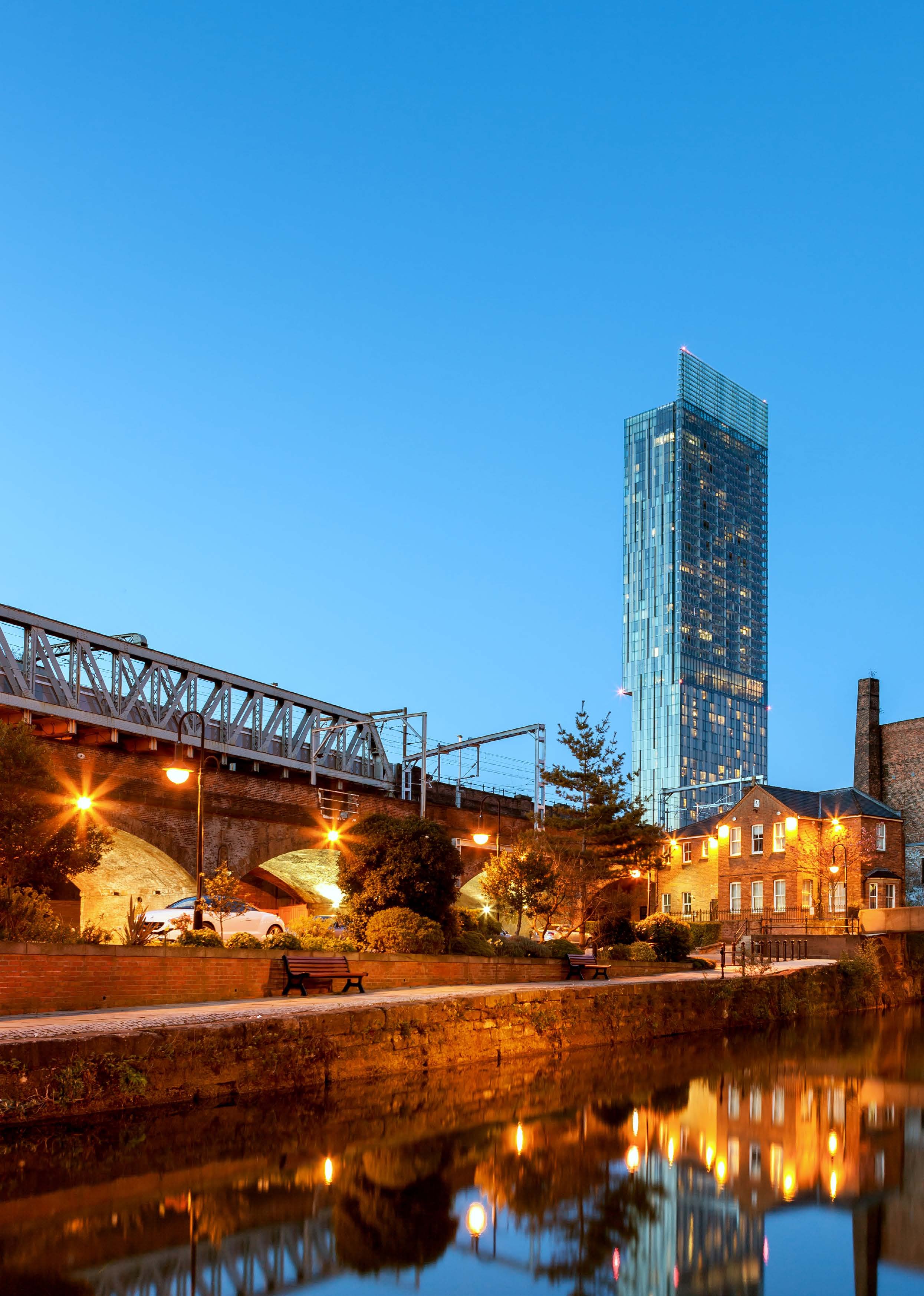 Evening view of a city canal with a railway bridge, historical brick building, and modern skyscraper under a blue sky.