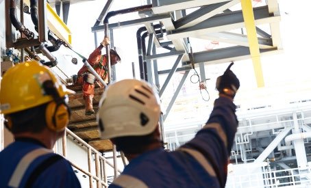 Industrial workers in hard hats and safety gear, one pointing, observing an overhead structure with pipes and scaffolding.