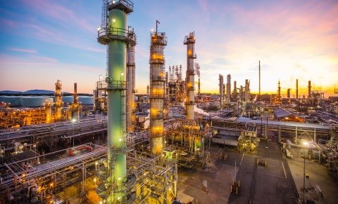 Aerial view of a large industrial facility or oil refinery at dusk, with illuminated towers and complex piping against a colorful sky.