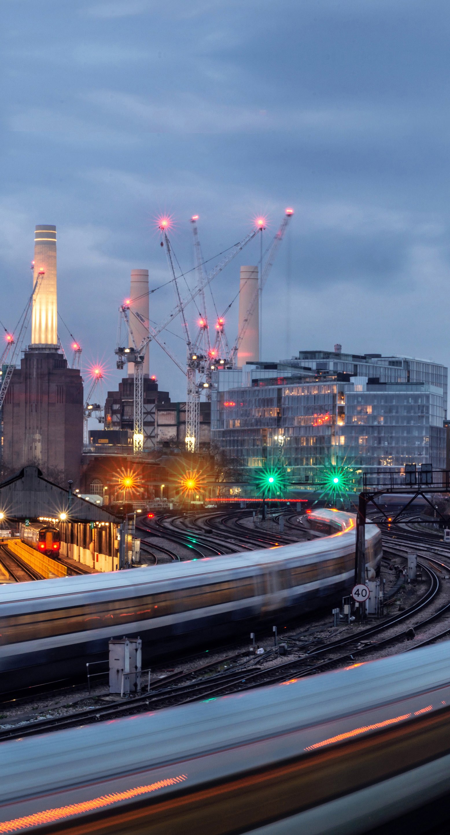Night scene of Battersea Power Station and surrounding railway tracks with blurred moving trains, cranes, and illuminated modern buildings.