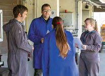 Four people in blue uniforms or lab coats discussing documents in an industrial or lab setting.