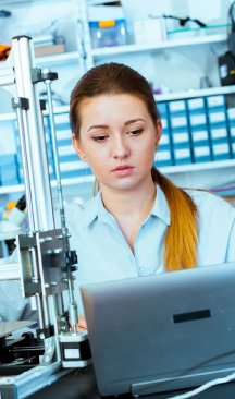 A woman with long hair looking intently at a laptop screen, positioned next to complex technical equipment in a laboratory or workshop setting.