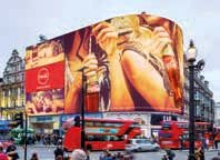 Busy urban street scene with a large digital billboard on a building, featuring advertisements and red double-decker buses below.
