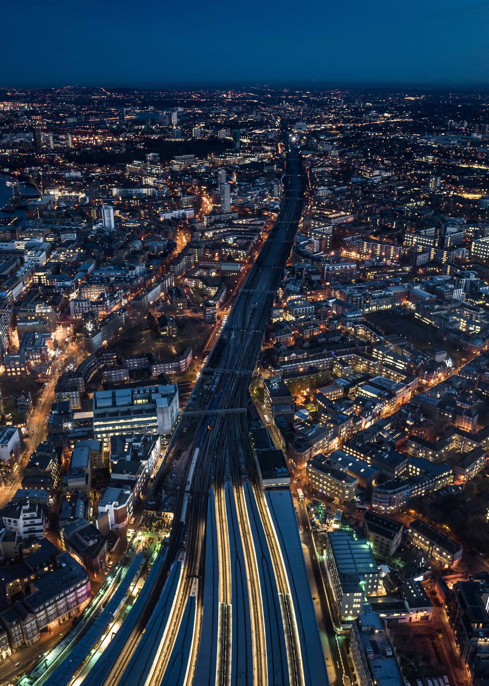 Aerial night view of a sprawling city with a prominent train station and railway lines cutting through illuminated buildings.