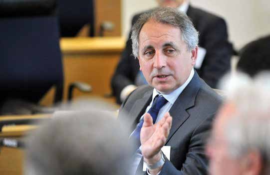 Man in a suit and tie speaking and gesturing with his right hand in a meeting room, with other participants blurred.