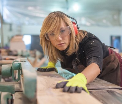 A woman wearing safety goggles and ear protection focuses while operating a woodworking machine, guiding a piece of wood with gloved hands.