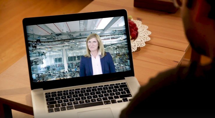 A person views a laptop screen showing a video call with a woman in a factory setting, suggesting a virtual tour or presentation.
