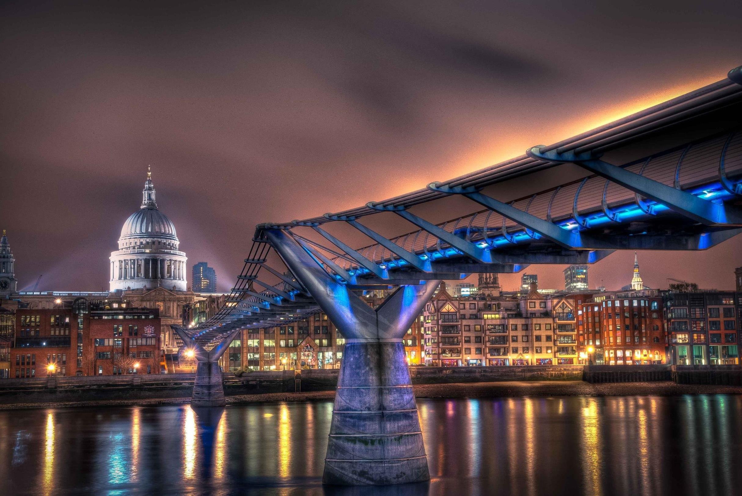 Nighttime view of the illuminated Millennium Bridge in London with St. Paul's Cathedral in the background, reflected in the River Thames.