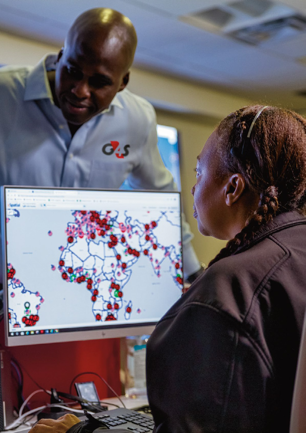 Two people, one pointing at a computer screen displaying a world map with numerous red and green data points across continents.