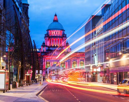 Illuminated Belfast City Hall at night, with long exposure car light trails on a busy street.
