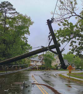 A utility pole has fallen across a wet road, with power lines visible. Trees and houses are in the background, suggesting storm damage or an accident.
