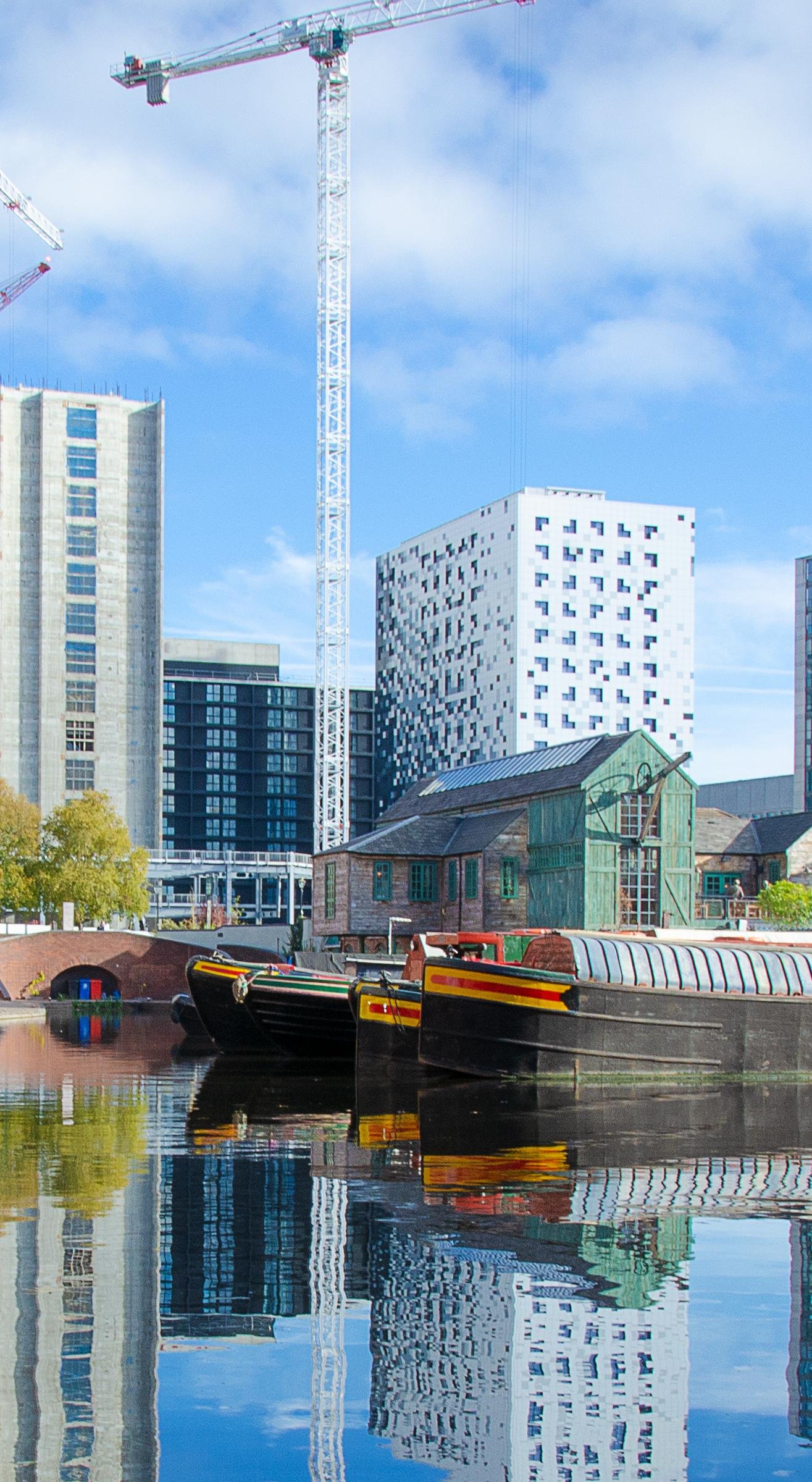 City canal scene with narrowboats, a historic green building, modern high-rise buildings, and construction cranes, all reflected in the calm water.
