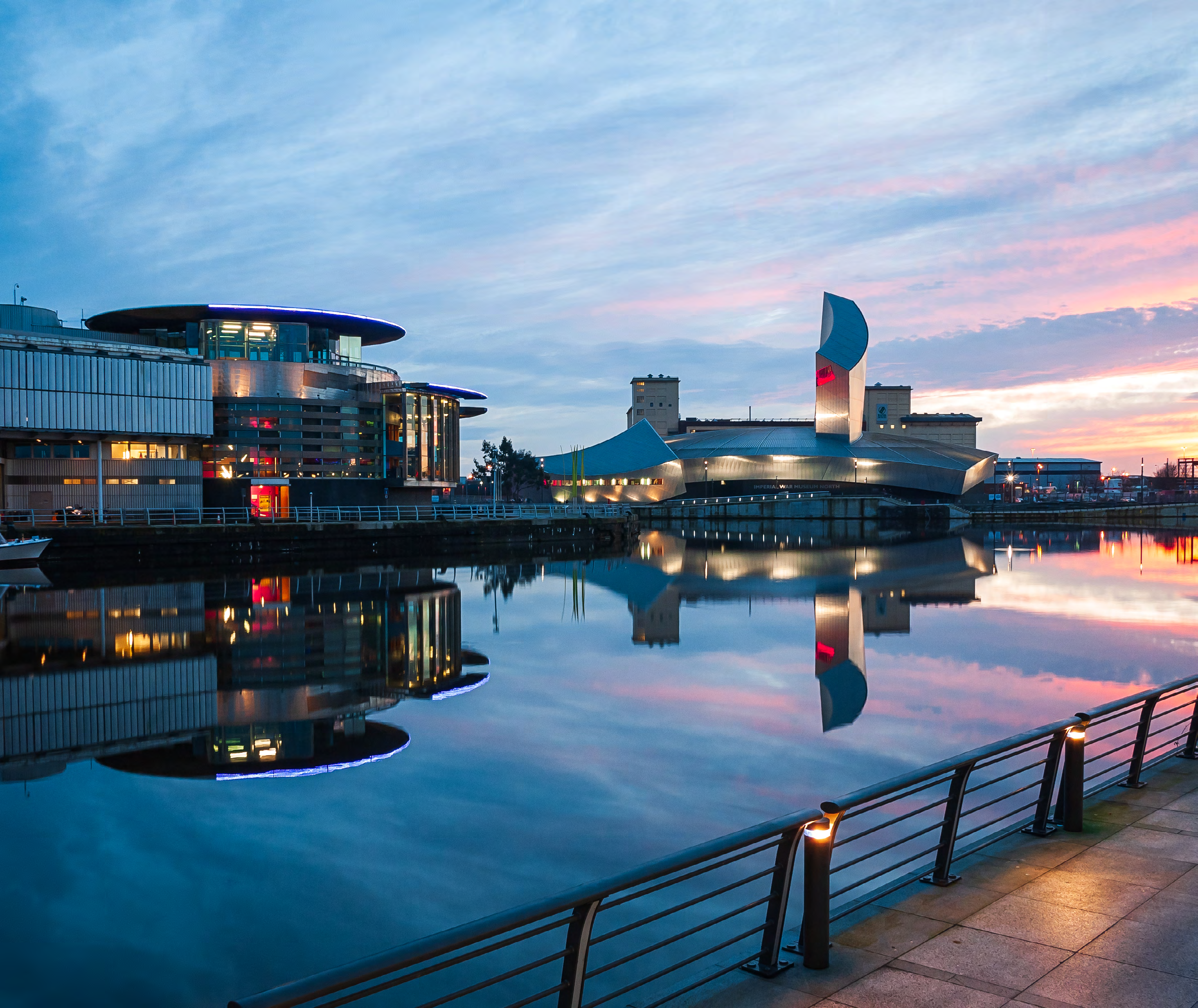 Scenic view of modern buildings reflecting in water at sunset or sunrise, with a colorful sky.
