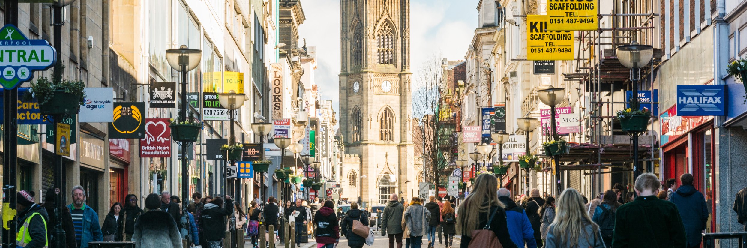 Bustling city street with pedestrians, shops, and a prominent clock tower in the distance.