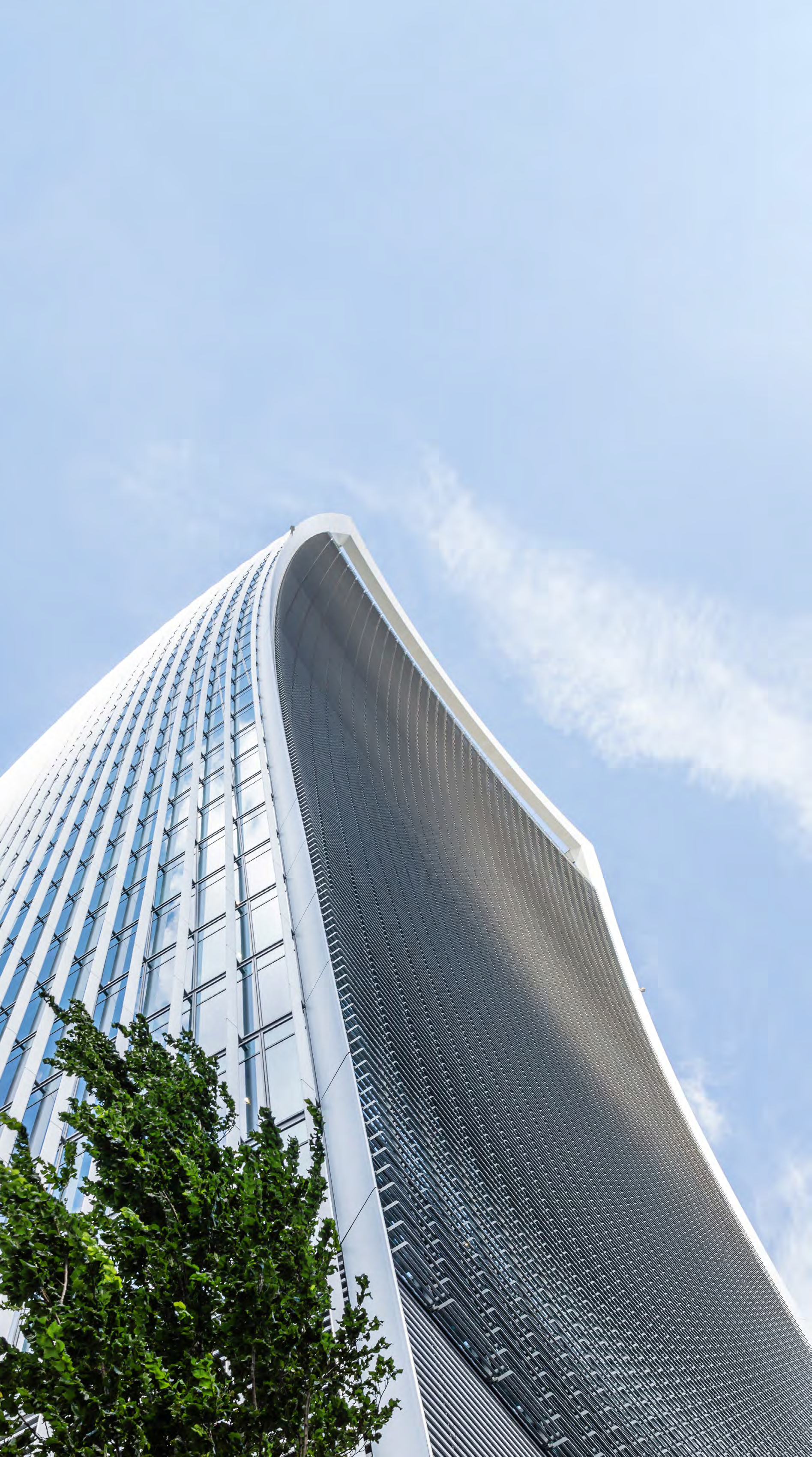 Low-angle view of a modern skyscraper with a curved, glass, and slatted facade, and a green tree in the foreground, against a blue sky.
