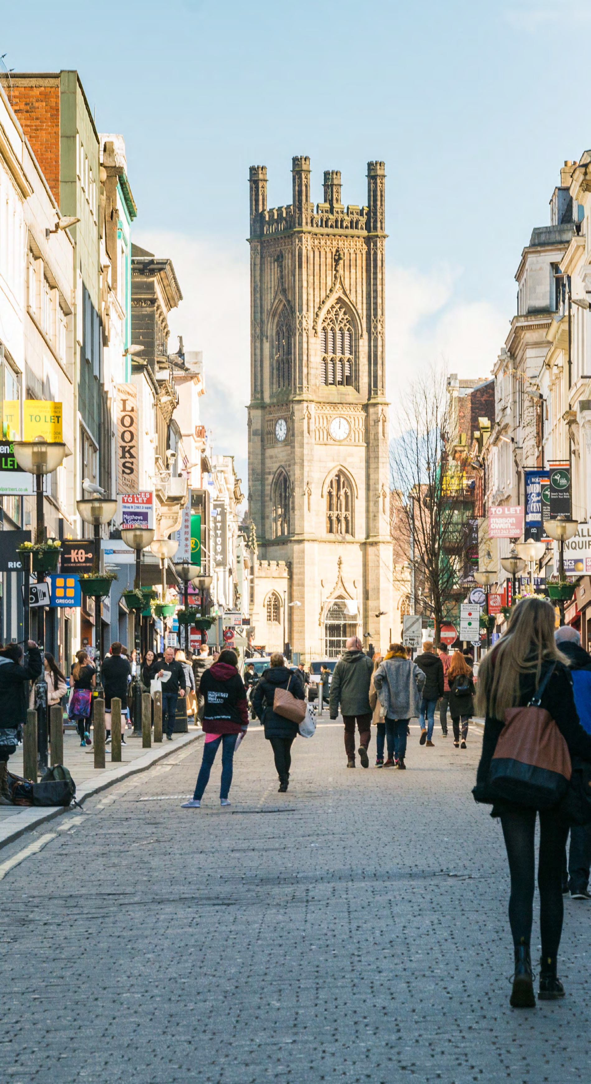 A busy cobbled street in a city, with people walking towards a prominent, tall Gothic church tower under a blue sky.