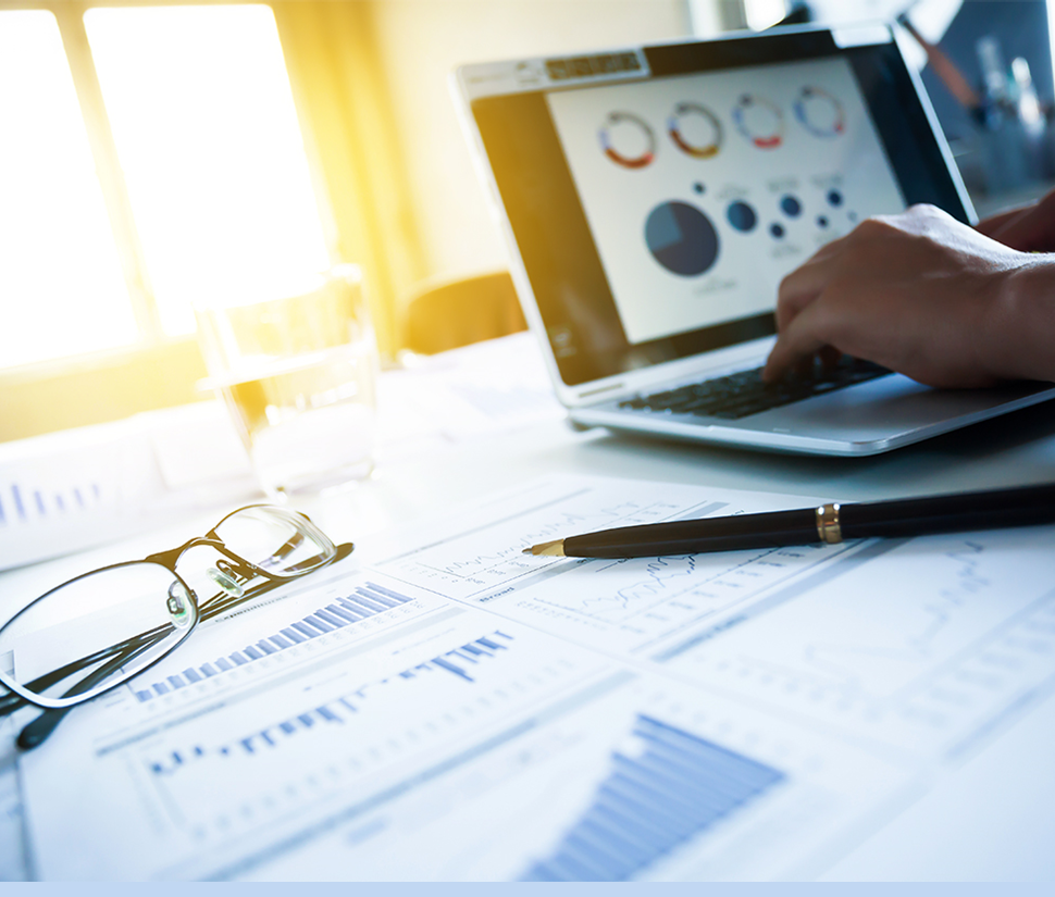 Person typing on a laptop displaying data charts, with papers showing graphs, glasses, and a pen on a sunlit desk, depicting financial analysis or business work.