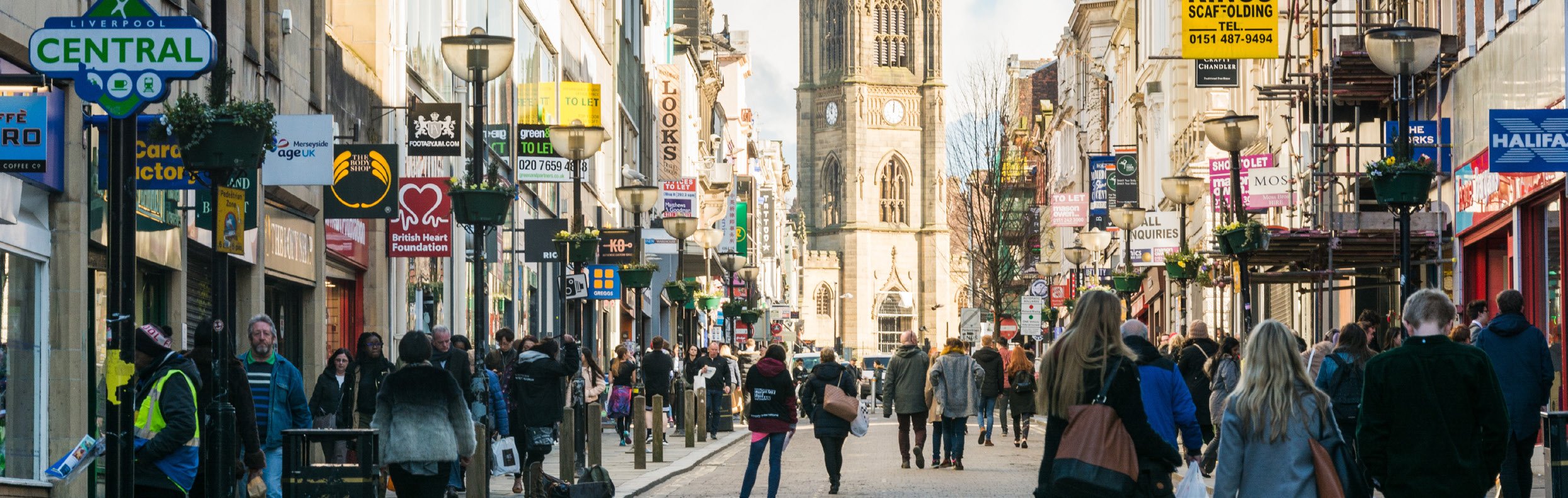 Bustling street scene in Liverpool city center with pedestrians, shops, and a prominent church tower. Signs for 'Liverpool Central' and various businesses are visible.