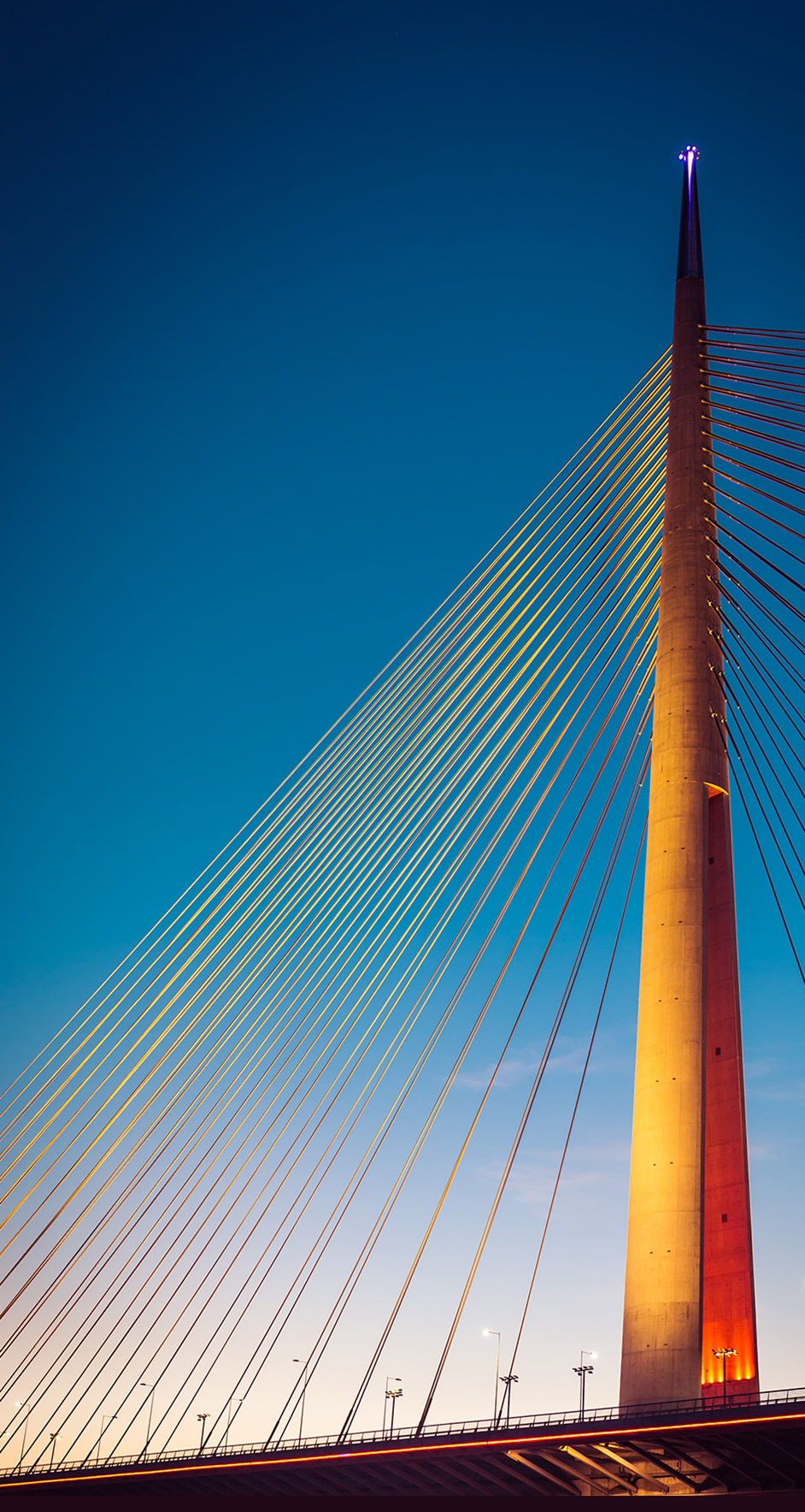 A modern cable-stayed bridge tower and cables lit by warm light against a deep blue evening sky.