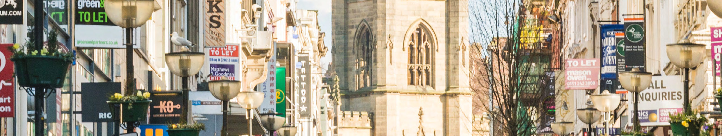 Bustling street scene with numerous commercial 'to let' signs, street lamps, planters, and a historic stone church tower.