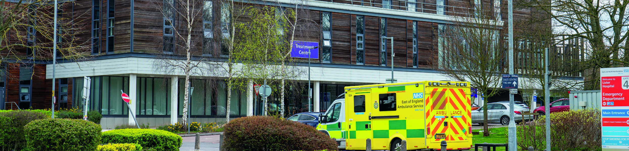 Exterior view of a hospital building with an NHS East of England ambulance parked in front. Signs for "Royal Lister Hospital" and "Emergency Department" are visible.