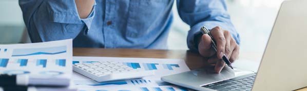 A person works at a desk with financial charts, graphs, a calculator, and a laptop, analyzing data.