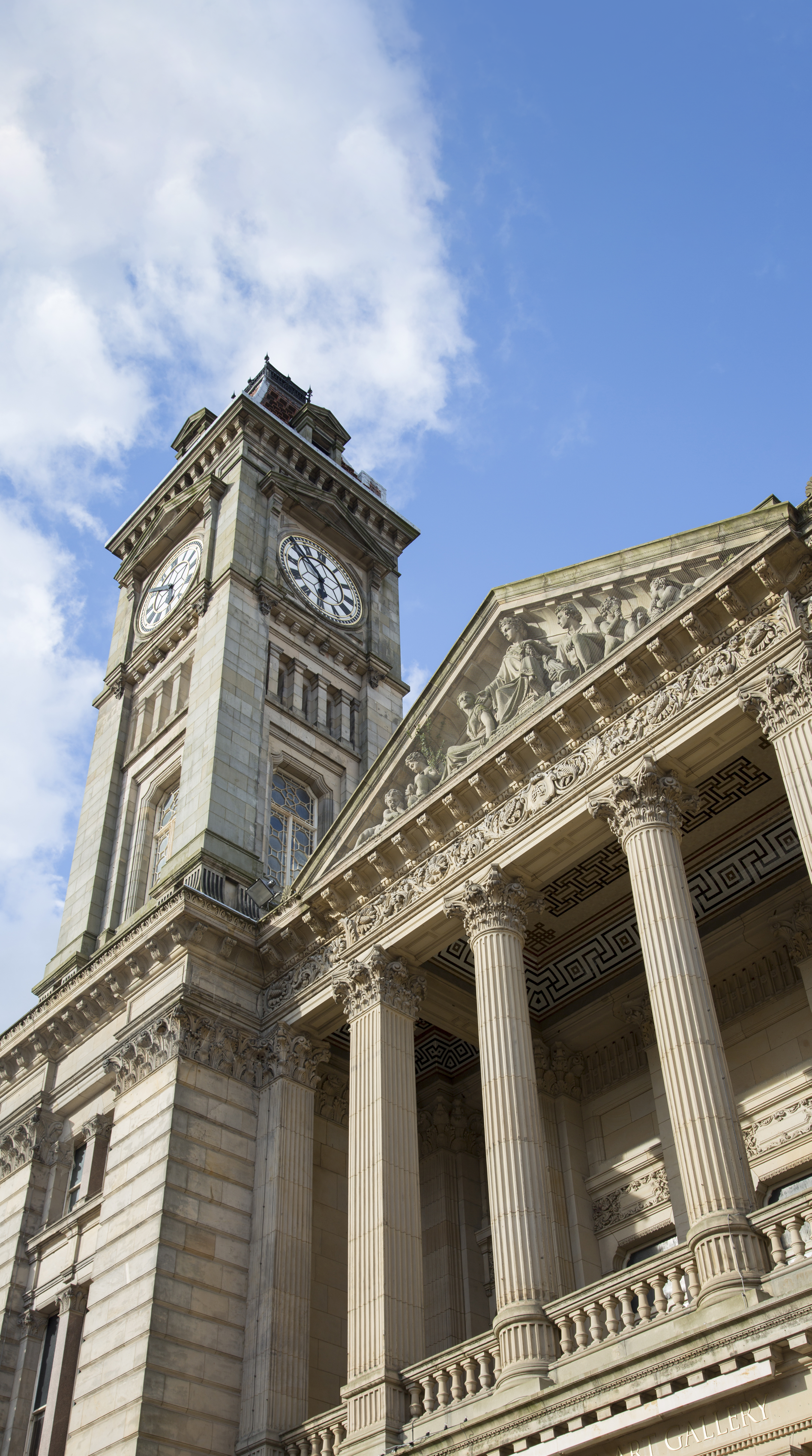 Low-angle view of a grand historic building with a prominent clock tower, classical columns, and sculptures on the pediment, set against a blue sky with clouds.