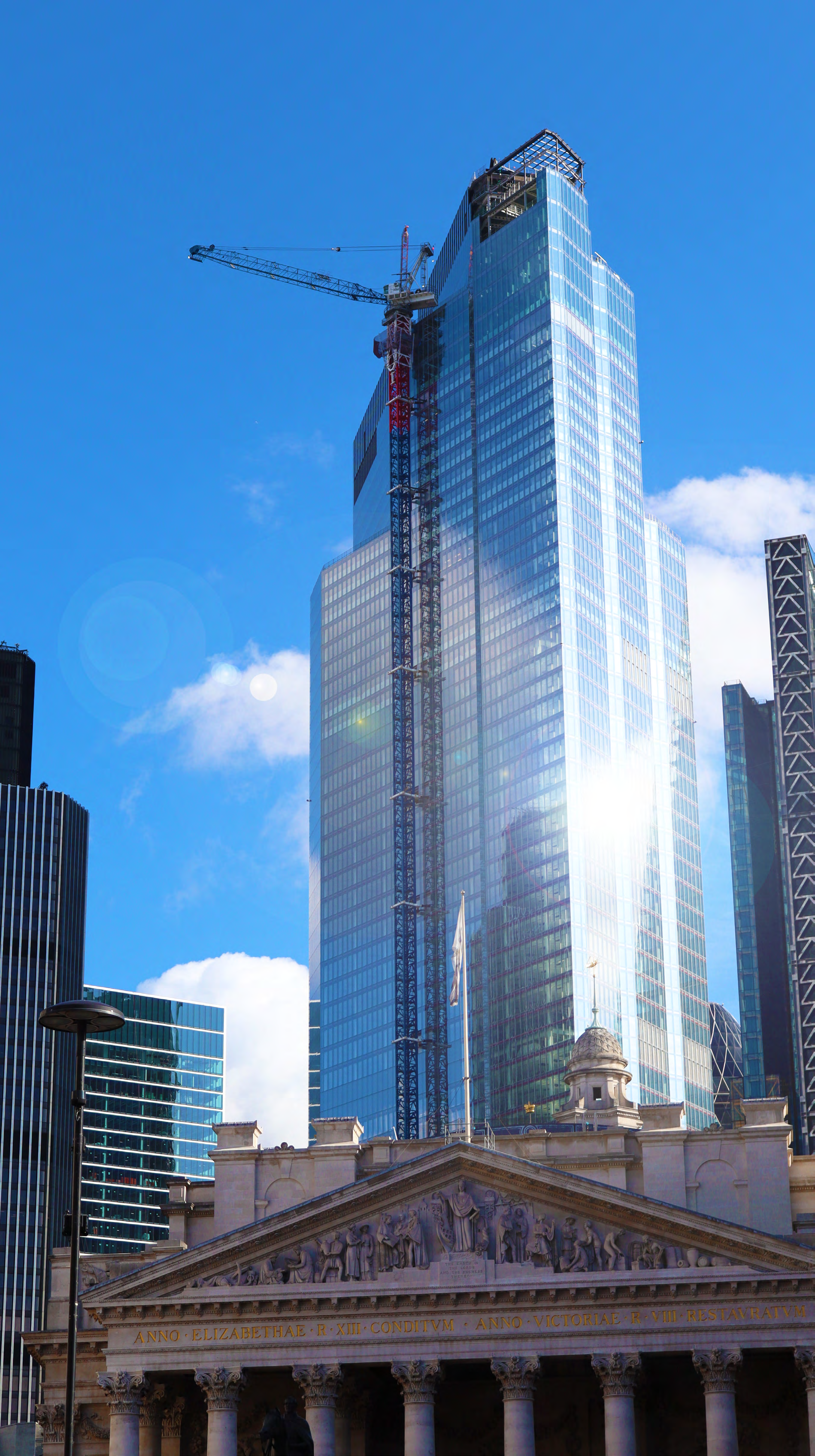 Urban landscape featuring a classical stone building with columns and a pediment in the foreground, juxtaposed with modern glass skyscrapers, one under construction with a crane, against a bright blue sky.