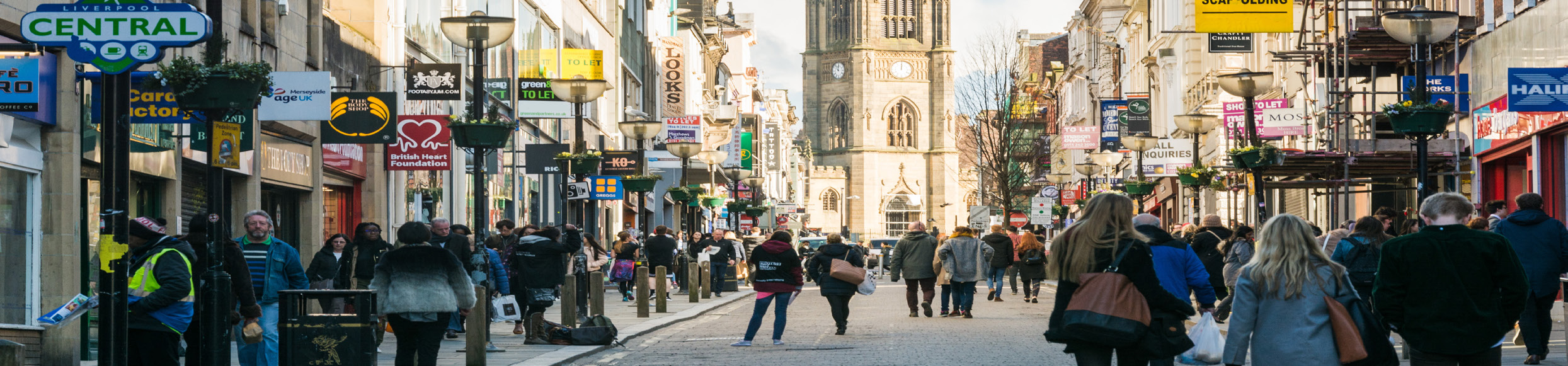 Bustling pedestrian street in Liverpool city center, with diverse shops, 'To Let' signs, and a prominent church tower.