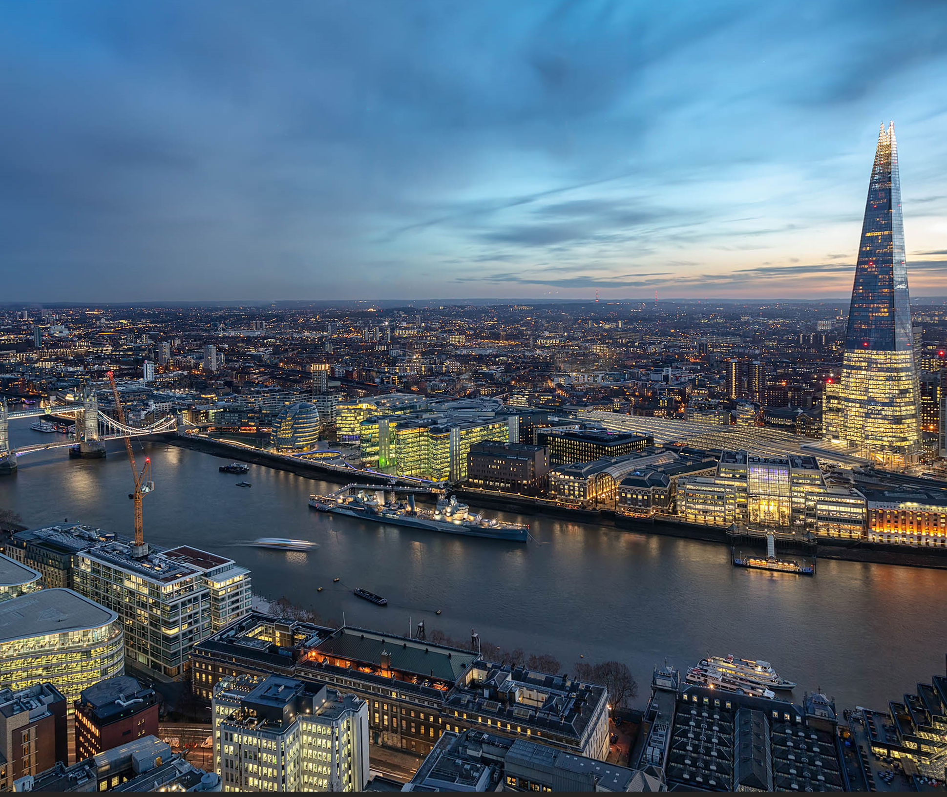 Aerial view of London at dusk, showing the River Thames, Tower Bridge, and numerous illuminated buildings including The Shard skyscraper.