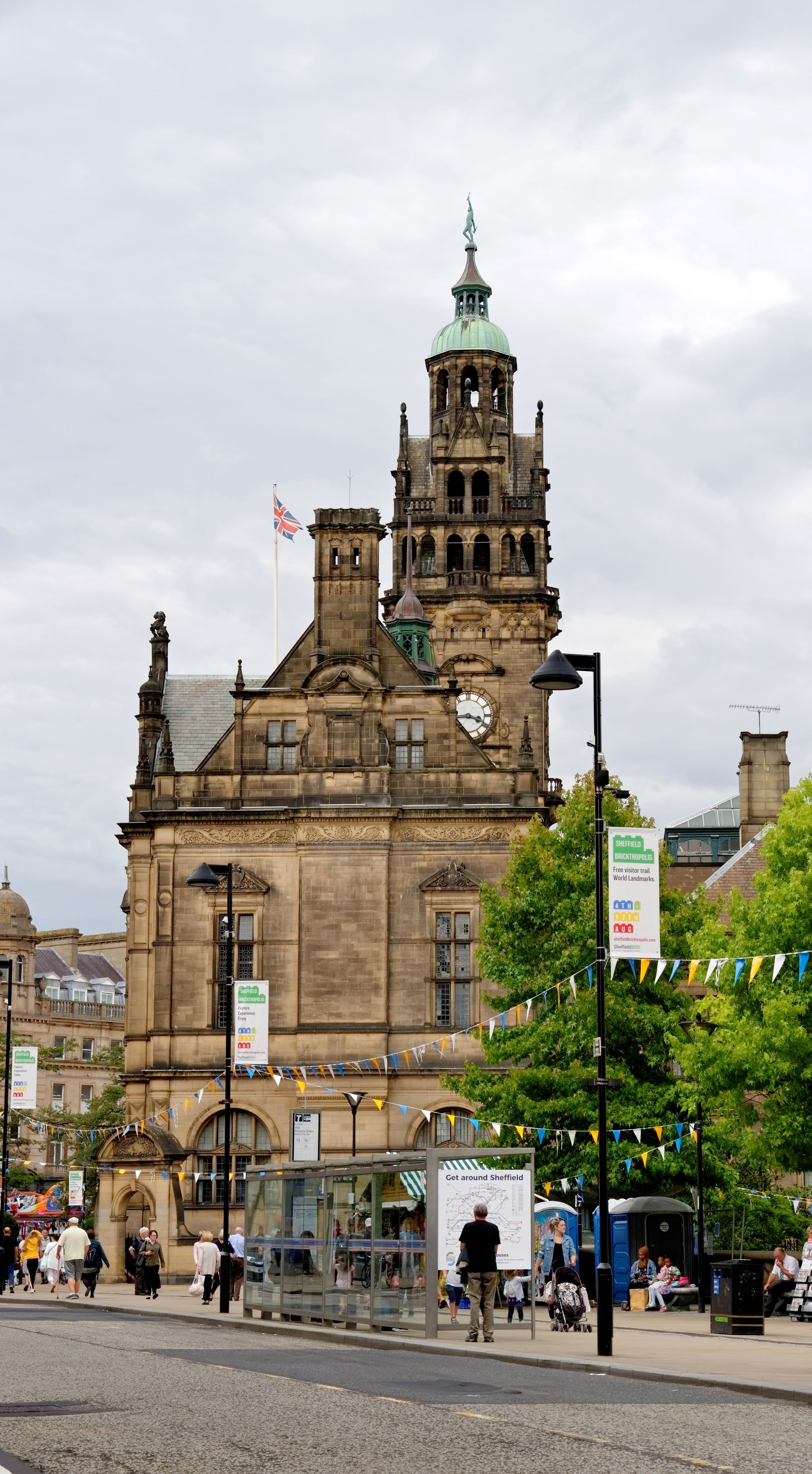 A grand stone building with a clock tower and a flag, people walk on the street in front. Likely Sheffield City Hall.