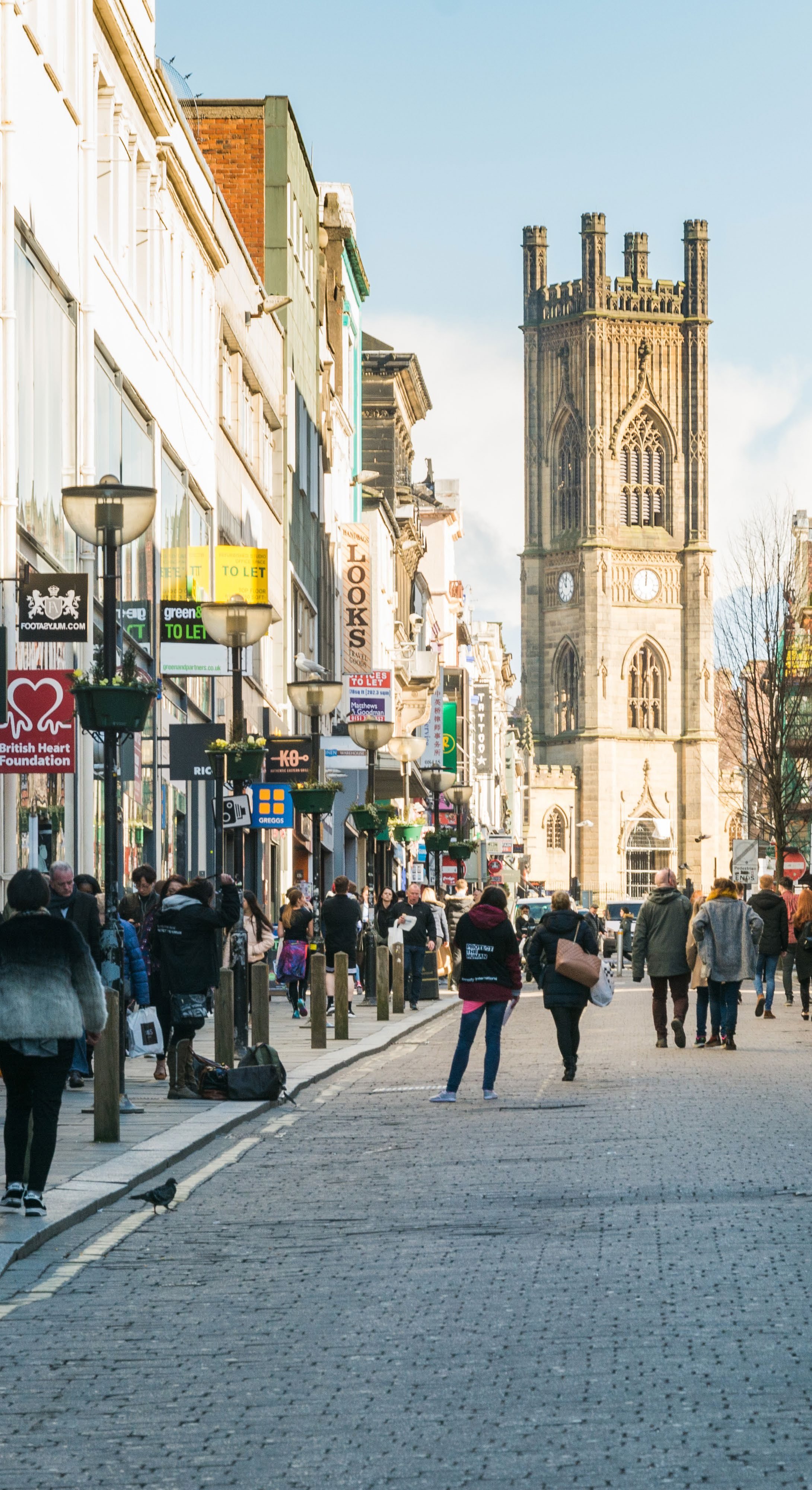 Street view of a city with shops lining a cobblestone street, people walking, and a tall gothic church tower visible in the background.