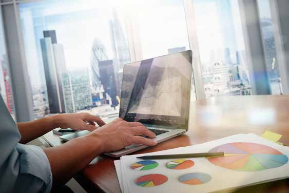 Hands typing on a laptop displaying a graph, with printed pie charts and documents on the desk, indicating data analysis.