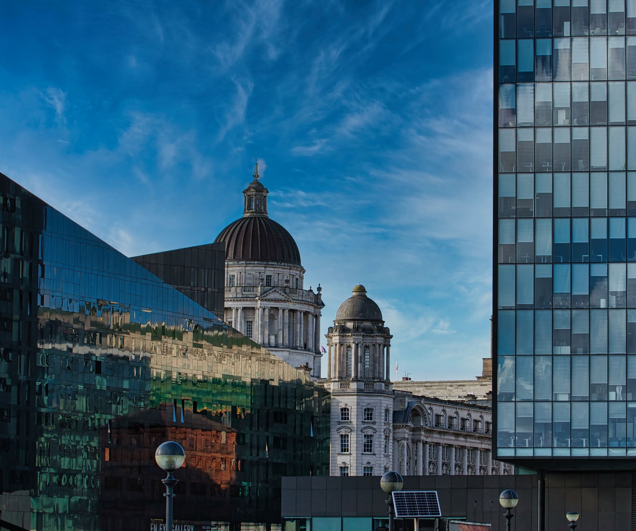 Diverse architecture with historic domed buildings reflected in the glass facades of modern skyscrapers under a blue sky.