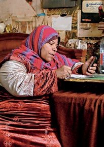 A woman wearing a colorful headscarf and patterned dress, sitting at a table and writing, with a phone nearby.