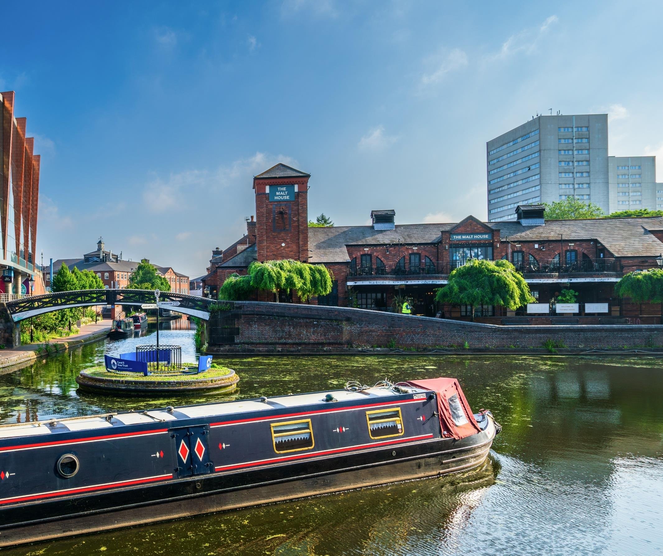 A canal scene in Birmingham, UK, with a narrowboat, brick buildings like "The Malt House," a bridge, and modern structures under a blue sky.