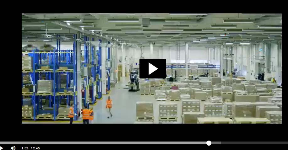 Screenshot of a video showing a vast warehouse or factory floor filled with stacked boxes, shelving, and workers