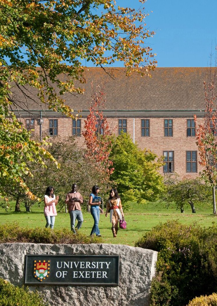 Students walk on the green campus of the University of Exeter, past a sign displaying the university's name and crest.