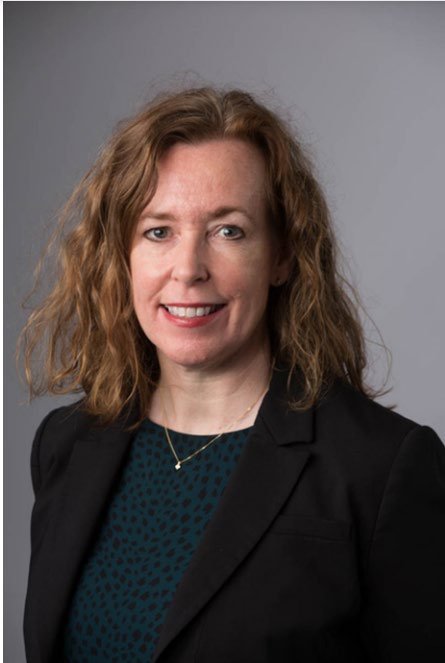 Headshot of a smiling woman with shoulder-length curly brown hair, wearing a dark jacket and patterned top, against a gray background.