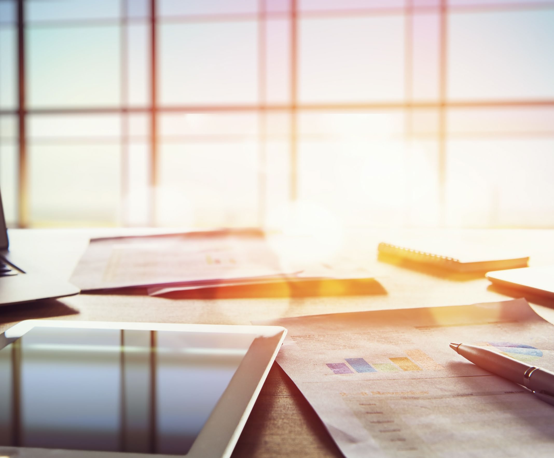 Office desk with a laptop, tablet, pen, and papers showing blurred charts, conveying a theme of business analysis.