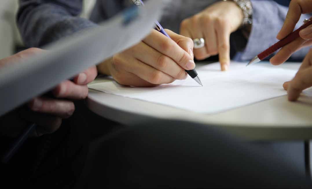 Close-up of hands working on a document, with one hand writing with a pen and another hand pointing, indicating collaboration.