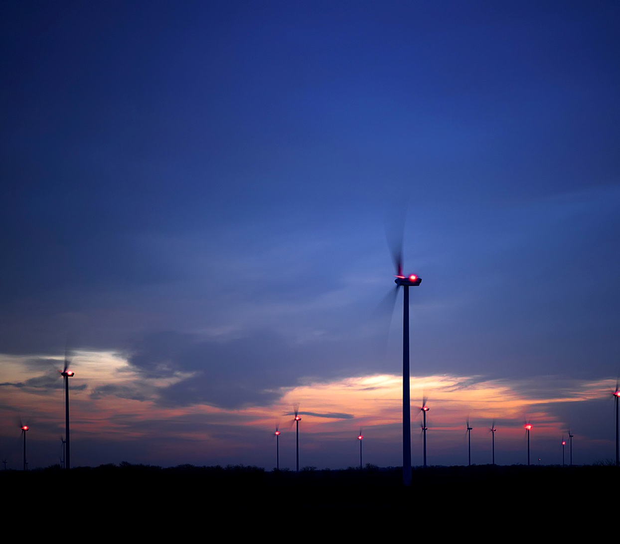 Wind turbines at dusk with red warning lights on the spinning blades, silhouetted against a dark blue and orange sky.