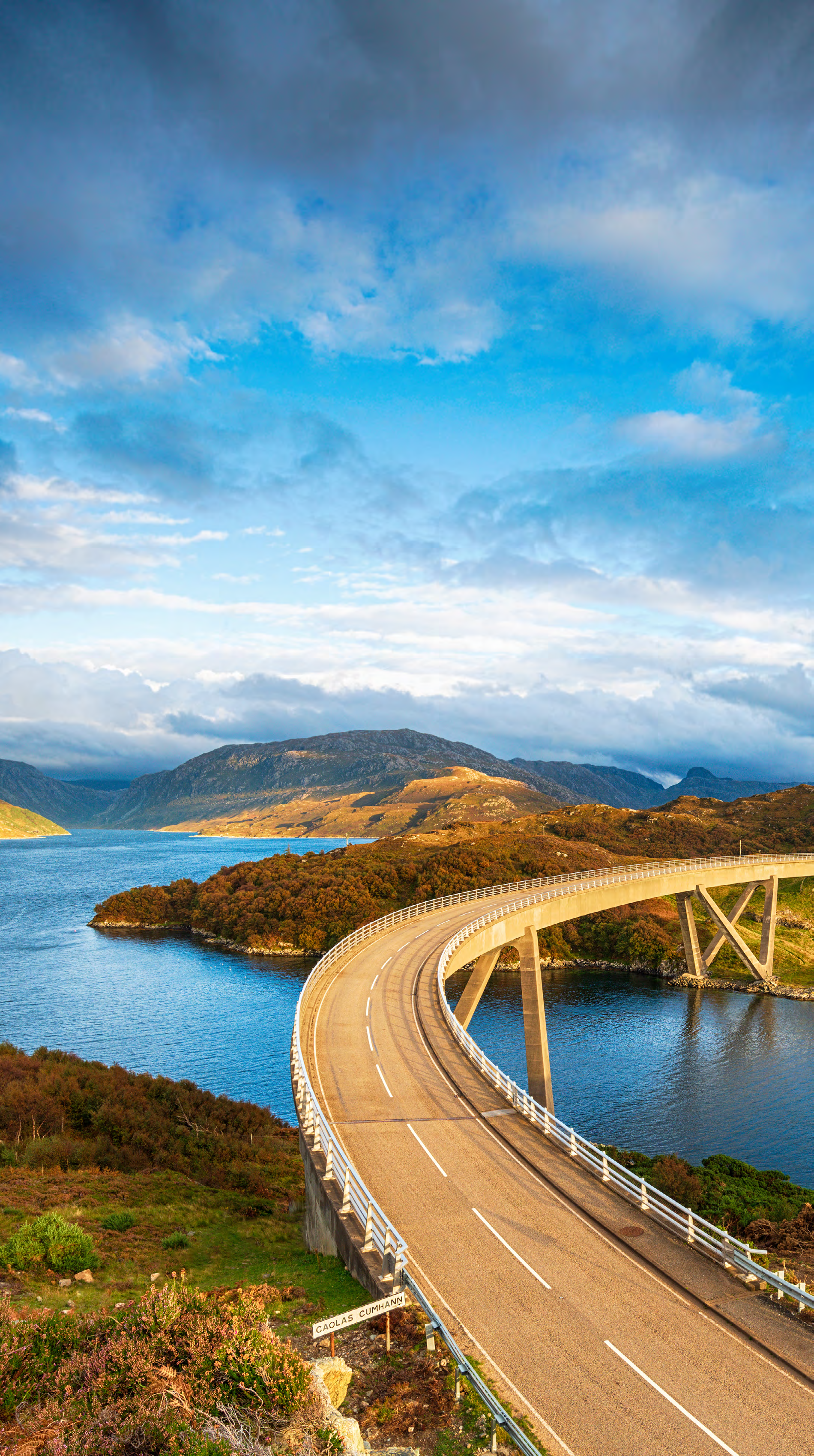 Curved concrete bridge spanning a body of water in a mountainous landscape under a cloudy sky.