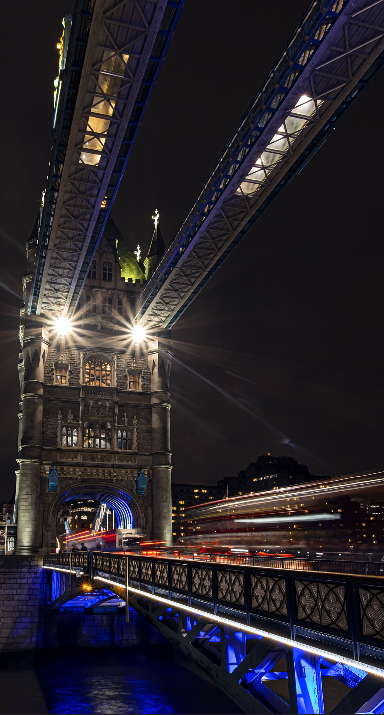 Night photo of Tower Bridge in London, illuminated, with red and white light trails from traffic moving across the bridge.