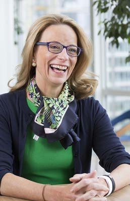 Portrait of a smiling woman with blonde hair and glasses, wearing a green top, blue cardigan, and patterned scarf.