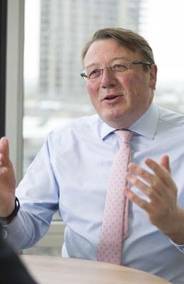 Portrait of a man with glasses, light blue shirt, and pink tie, gesturing with both hands while speaking.
