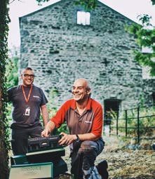 Two smiling men in workwear holding a toolbox, standing outdoors in front of an old stone building.