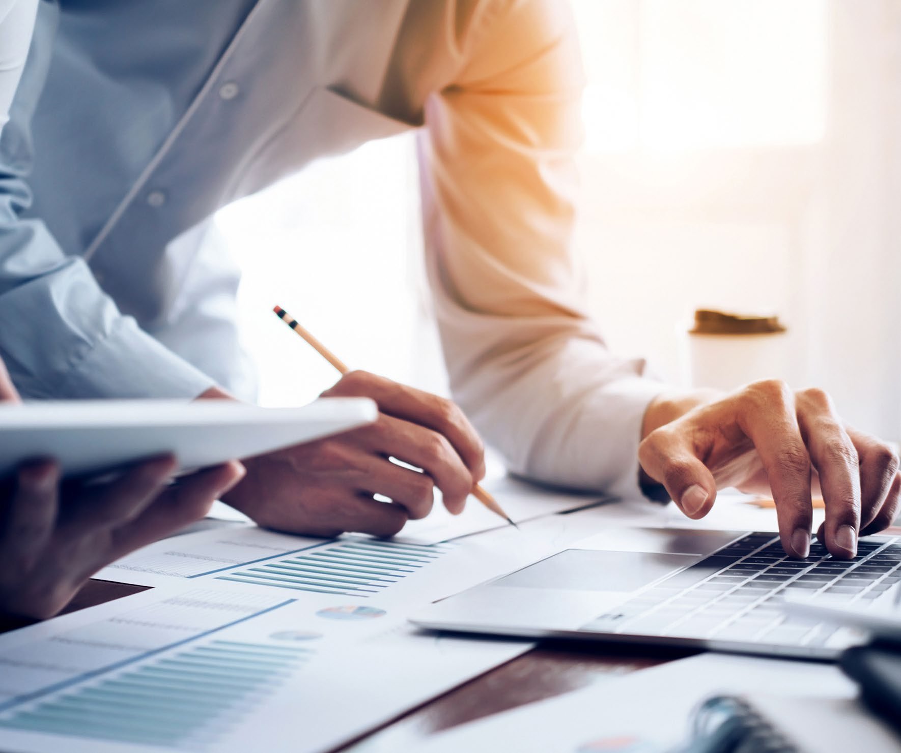 Close-up of hands collaborating around a table, reviewing financial charts and data on documents, a laptop, and a tablet.