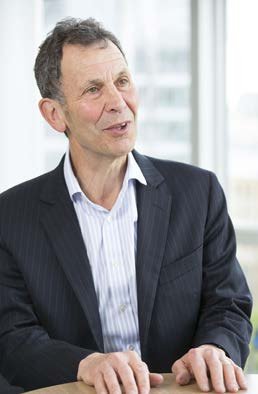 A man with dark hair, wearing a dark pinstripe suit and a striped shirt, speaks while resting his hands on a table.