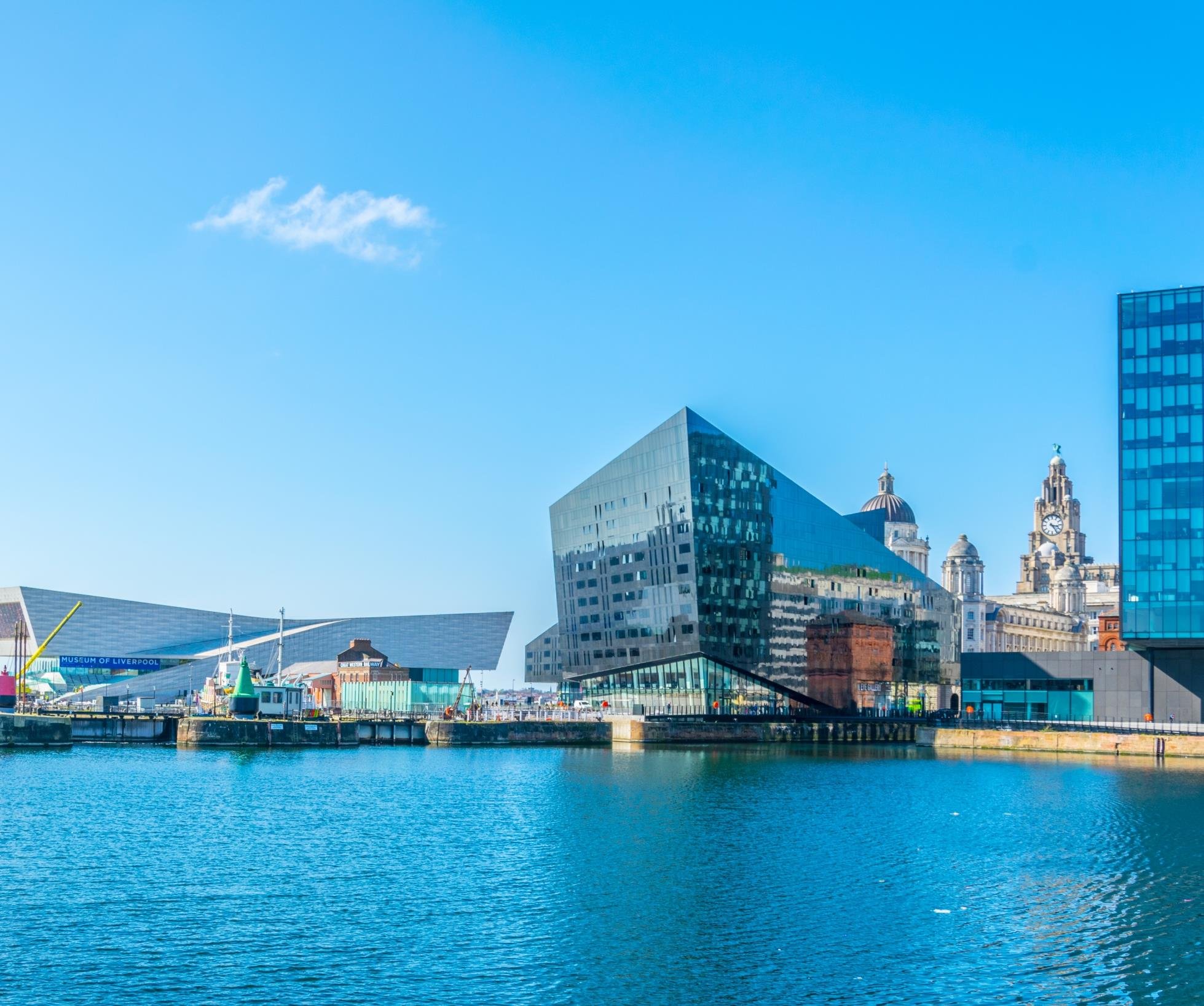 Panoramic view of Liverpool waterfront, featuring the Museum of Liverpool, modern glass buildings, and the historic Royal Liver Building with its iconic clock tower against a blue sky.